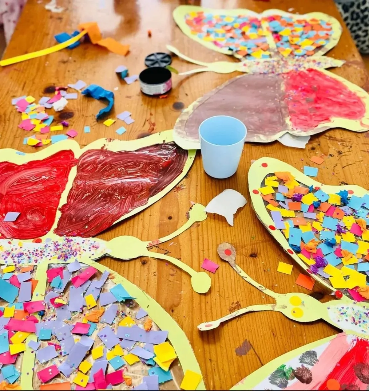 Mesa de trabajo llena de mariposas de papel, pinturas y materiales de collage